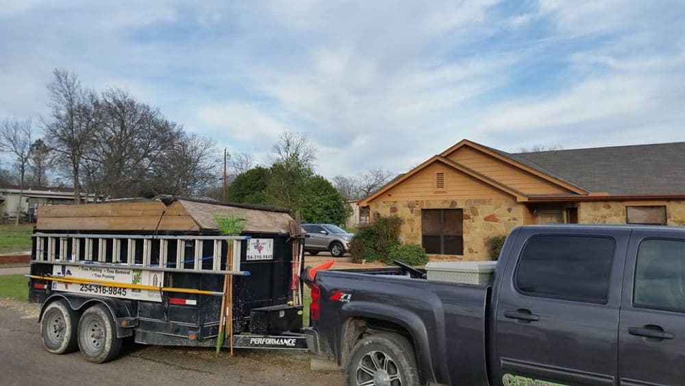 Pickup truck with trailer carrying debris in front of a single-story house on a cloudy day.