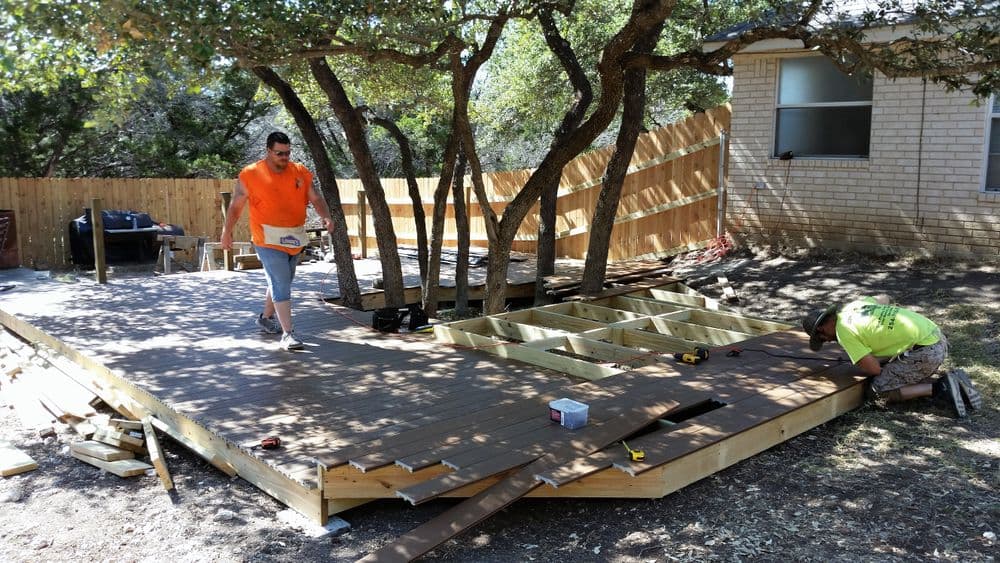 Workers constructing a wooden deck in a yard with trees and a house in the background.