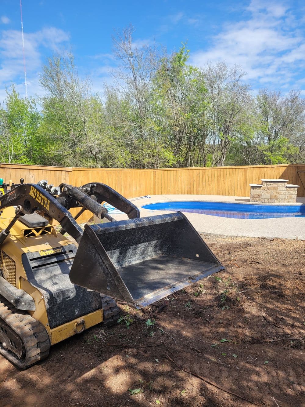 Skid steer loader preparing landscape near a newly constructed pool and wooden fence.