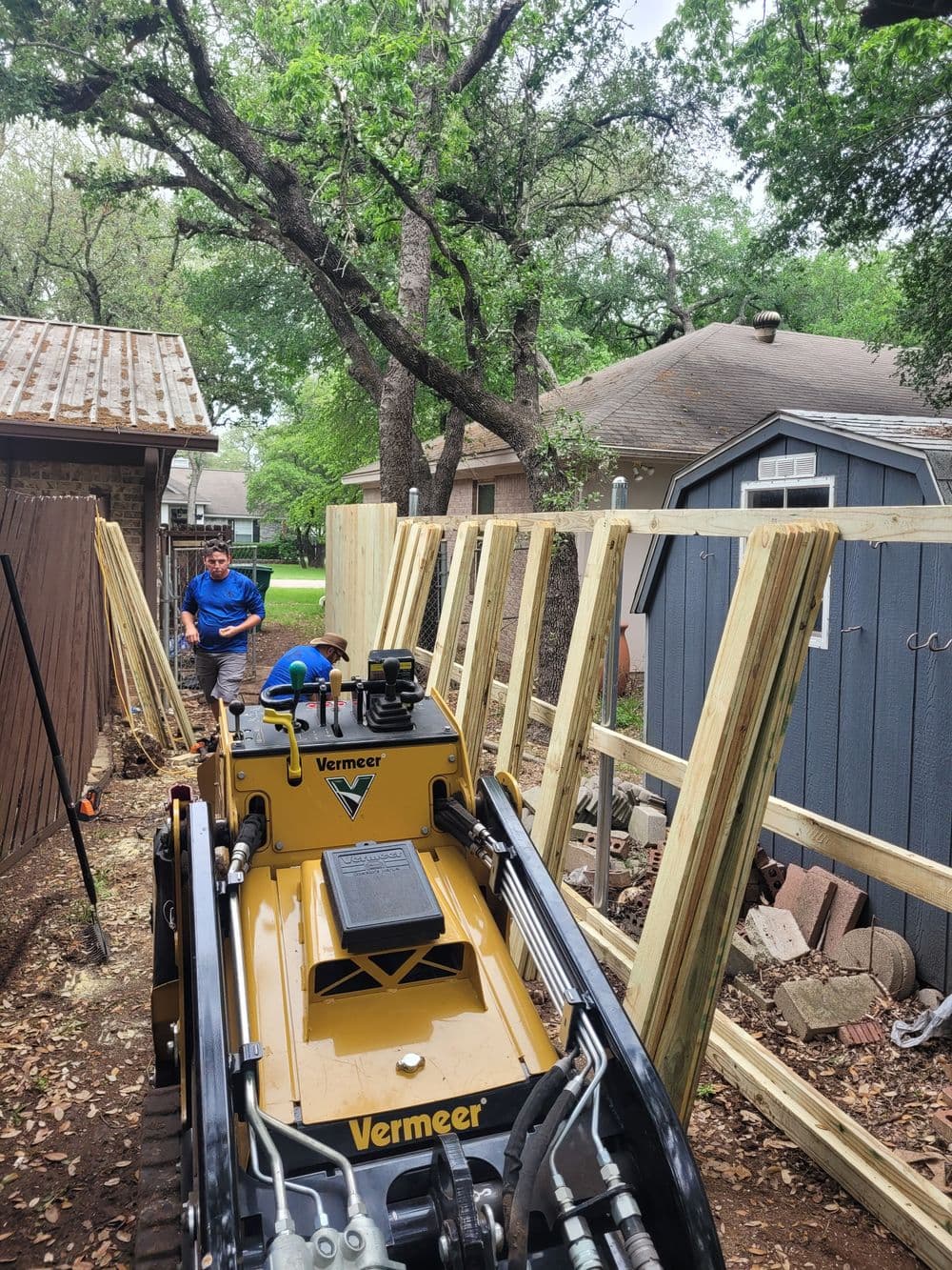 Vermeer machine in yard with worker installing wooden fence panels among trees.